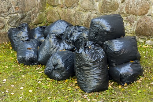 A collection of nine black rubbish bags made of plastic, filled with waste and tied at the top, situated on a grassy area in front of a stone wall. The bags vary slightly in shape and size, some appearing more full and rounded, while others are more squashed or flattened. The bags are placed directly on the grass, which is interspersed with small scattered leaves and pebbles. The stone wall behind comprises irregularly shaped, weathered stones with visible moss and lichen growth, providing a textured, natural backdrop. The scene is outdoors, likely during the daytime, with diffuse natural light highlighting the sheen of the plastic bags and the texture of the grassy ground and stone wall. This setup may be part of a waste collection or rubbish removal process, aligning with services offered by Commercial Waste Welwyn in the local area near Welwyn, Hertfordshire.