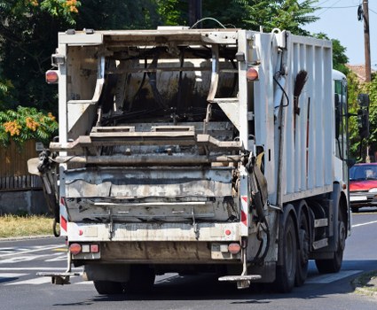 A rear view of a large industrial rubbish collection vehicle, painted in light grey, operating on a rural street. The rear loading compartment is open, revealing a wide, heavy-duty steel hopper with visible wear and grime, as well as shredding or crushing mechanisms inside. The vehicle has a sturdy, rectangular metal body with textured surfaces, some rust and dirt marks, and red and white reflective warning strips along the lower edges. The back doors are slightly ajar, with hydraulic mechanisms visible on the sides for tilting or lifting. The vehicle is positioned on a paved local road, with trees, utility poles, and a parked car visible in the background, indicating a residential or small commercial area typical of Welwyn or surrounding regions. Bright daylight illuminates the scene, emphasizing the weathered surface of the waste truck, which is used by commercial waste removal services like Commercial Waste Welwyn for rubbish collection.