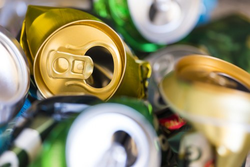 Close-up image of multiple aluminium beverage cans, predominantly gold and silver in color, with some green and blue cans visible in the background. The cans are crumpled and stacked irregularly on a surface, likely representing waste ready for collection. The tops of the cans show pull tabs, with some open and others closed. The textured surfaces reflect light, emphasizing their metallic finishes. The surrounding environment appears to be a collection area or a waste disposal site, possibly on a paved driveway or outdoor space near a building in Welwyn. The scene captures the clutter of recyclable rubbish typical of waste removal services such as those offered by Commercial Waste Welwyn, emphasizing the importance of proper disposal and recycling of aluminium cans in the local area.